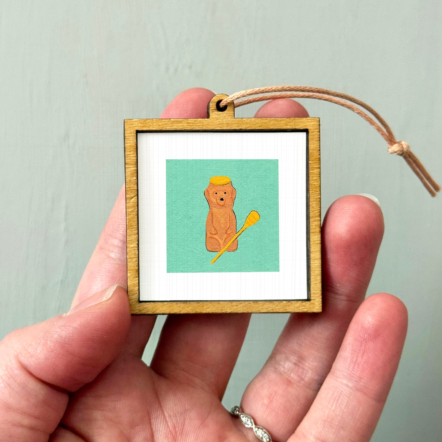 A person's hand holding a small wooden frame containing a picture of a brown bear with a yellow hat.