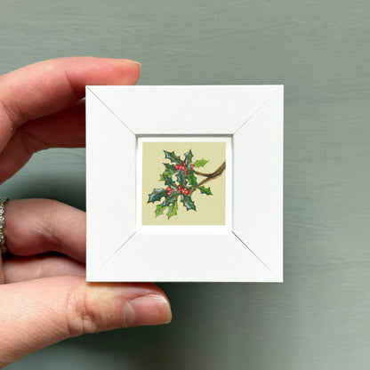 A hand holding a small white square frame containing a painting of a holly branch with berries.