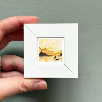 A hand is holding a small white square frame containing a watercolor painting of a landscape with mountains and a boat on a lake.