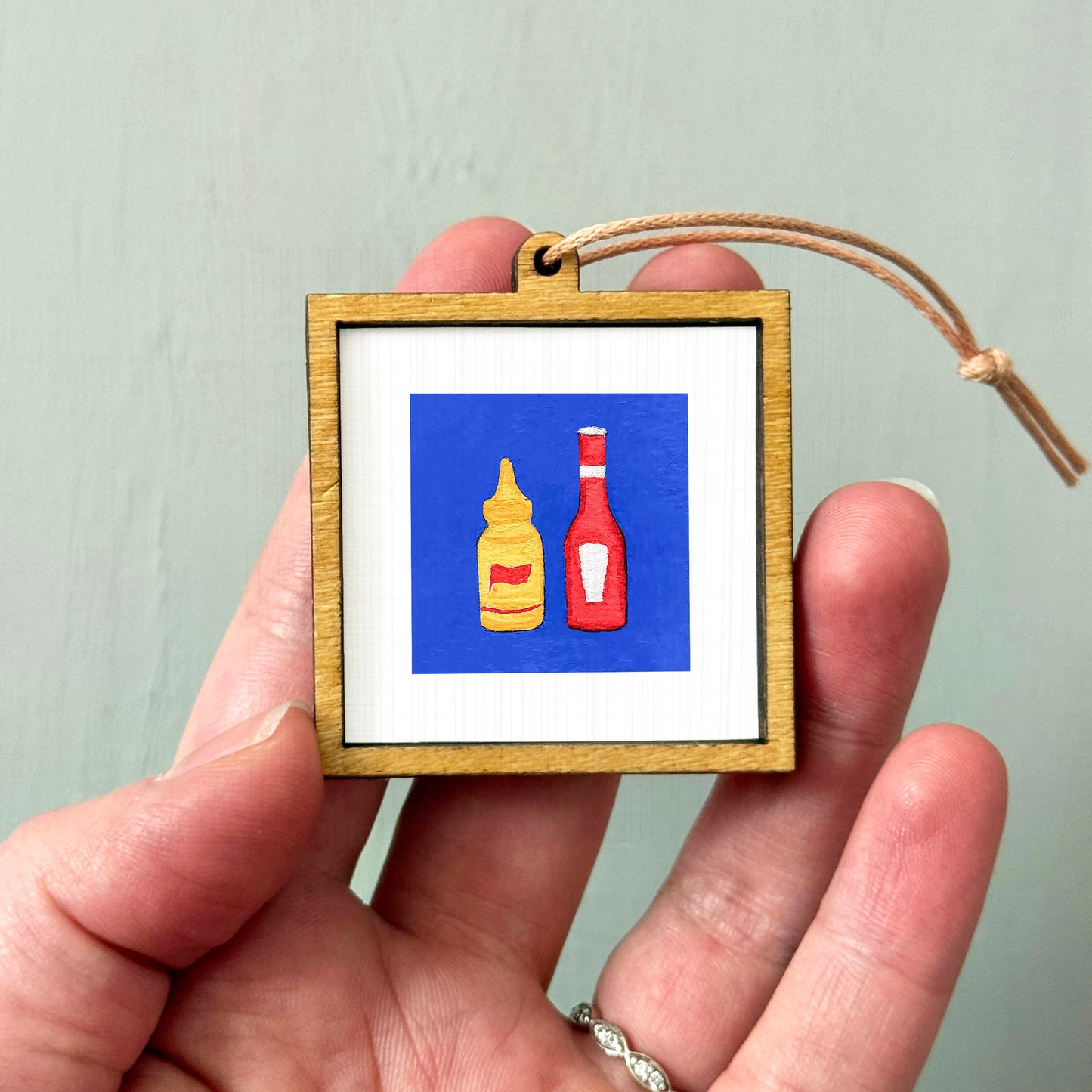 A person's hand holding a small wooden frame with a picture of two bottles of condiments inside it.
