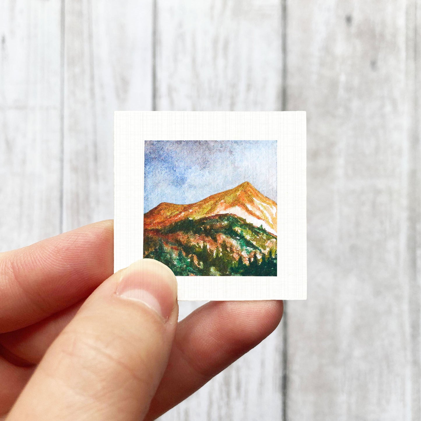 A hand is holding a small square-shaped painting depicting a mountain landscape with a colorful sky and trees.