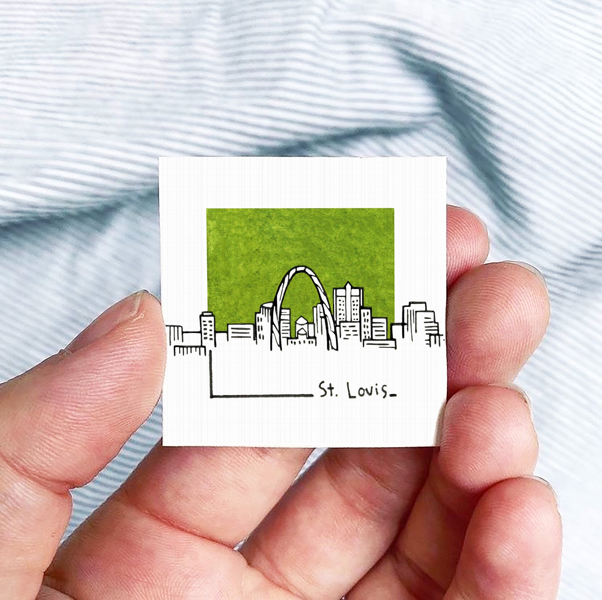 A hand holding a small square-shaped paper with a drawing of St. Louis' skyline.