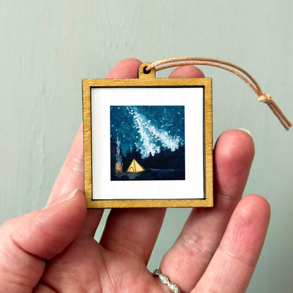 A hand holding a small wooden frame containing a painting of a starry night sky with a tent in the foreground.