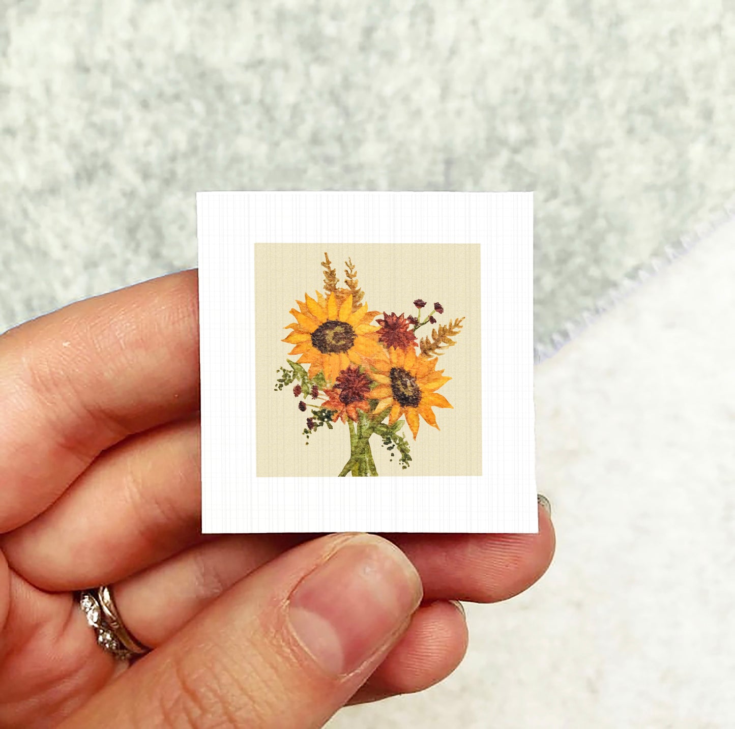 A hand holding a small square card with a vibrant floral design of sunflowers.