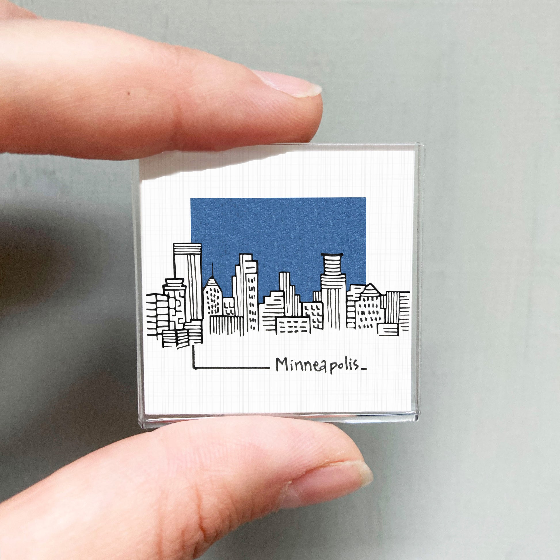 A hand holding a small square-shaped object with a drawing of a city skyline and the text " Minneapolis."