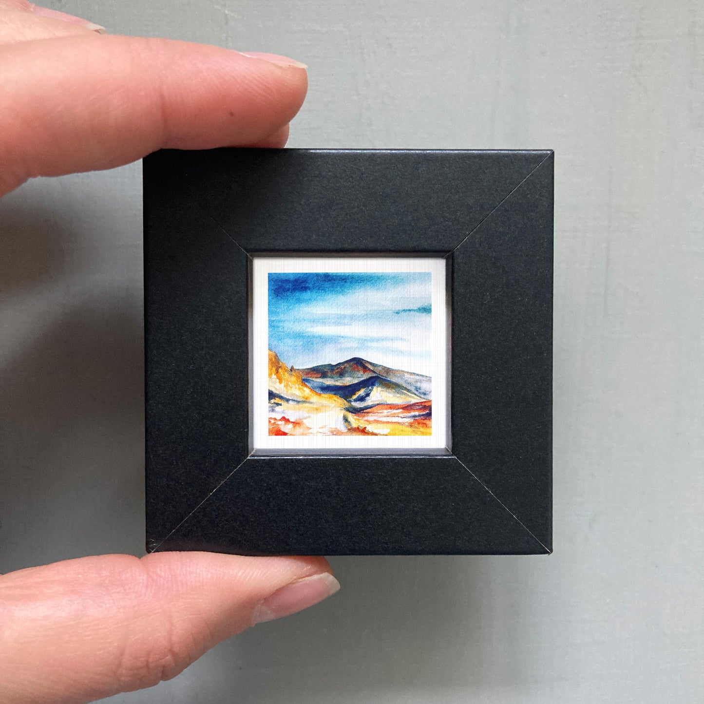 A person's hand holding a small, square-shaped black frame containing a watercolor painting of a mountain landscape.