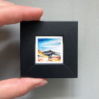A person's hand holding a small, square-shaped black frame containing a watercolor painting of a mountain landscape.