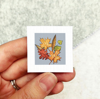 A hand holding a small square card with a colorful autumn leaf illustration on it.
