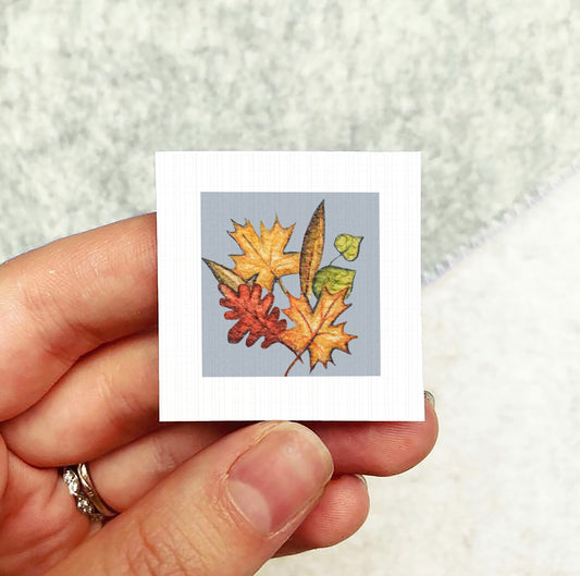A hand holding a small square card with a colorful autumn leaf illustration on it.