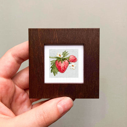 A hand is holding a small wooden frame containing a framed illustration of a strawberry with white flowers.