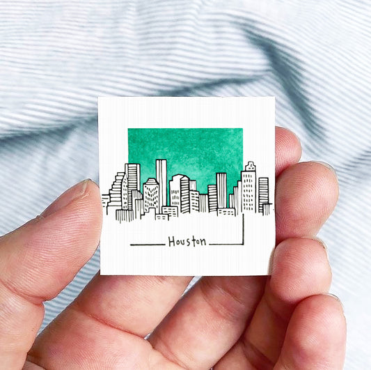 A hand is holding a small square-shaped object with a drawing of a city skyline and the word "Houston" written on it.