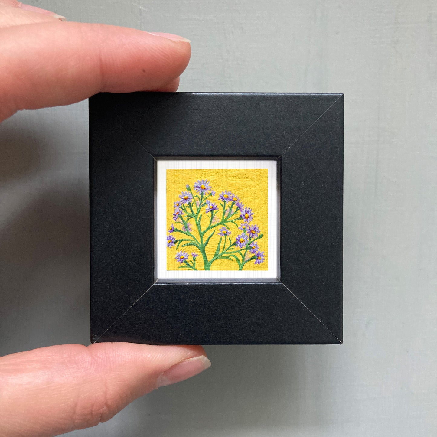 A hand is holding a small black frame containing a vibrant yellow and green floral painting.