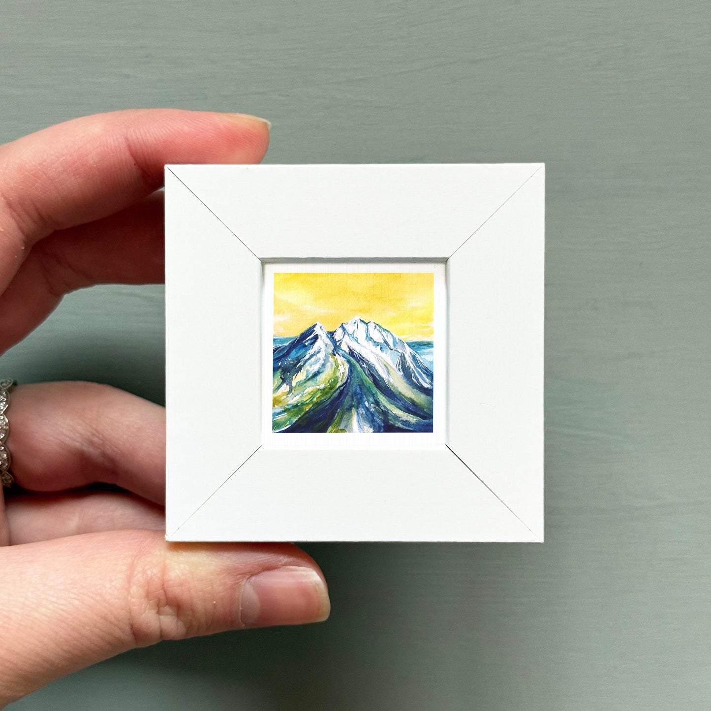 A hand is holding a small white picture frame containing a painting of a mountain landscape.