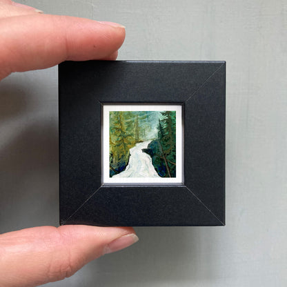 A person's hand holding a small black frame containing a framed painting of a winding river surrounded by trees.