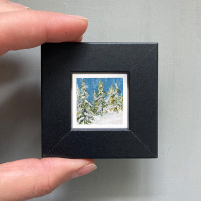 A person's hand holding a small black picture frame containing a painting of snow-covered pine trees.