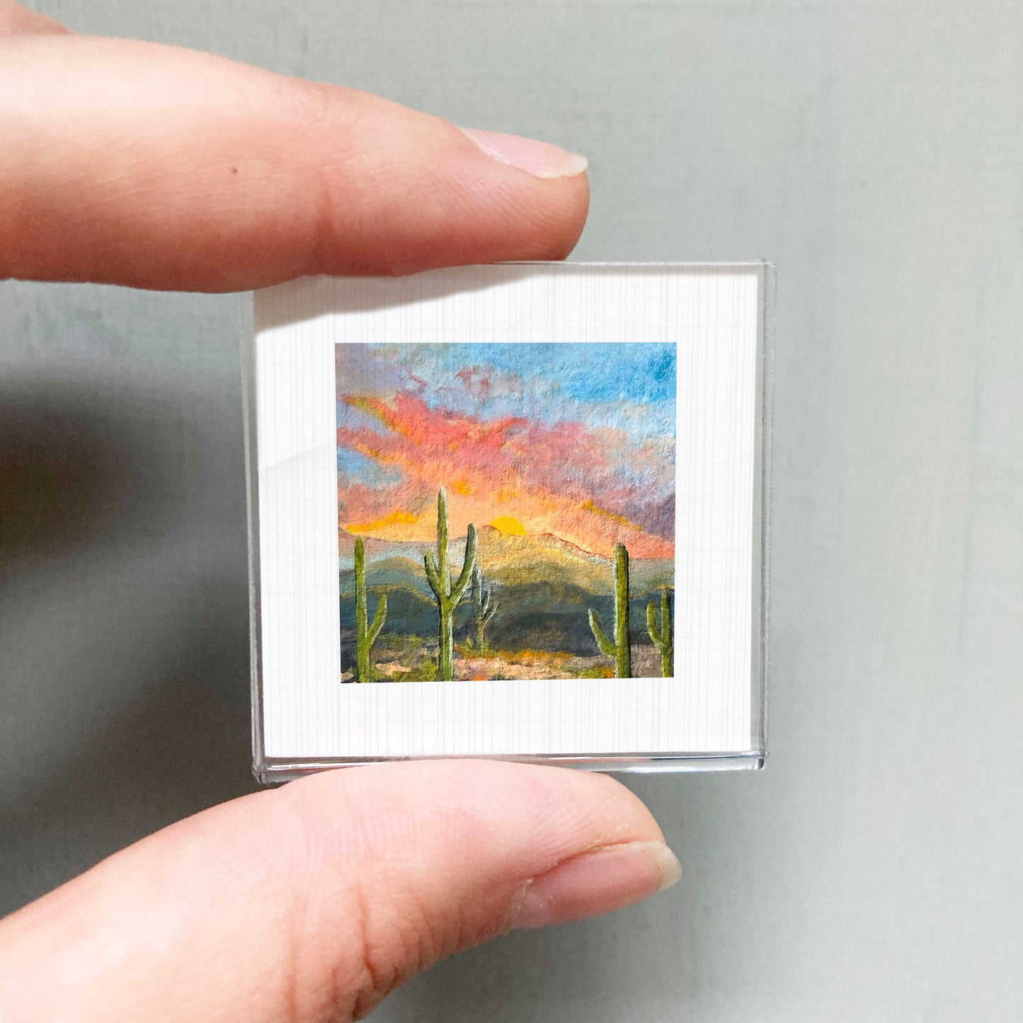 A person's hand holding a small square picture of a desert landscape with cacti and a colorful sunset sky.