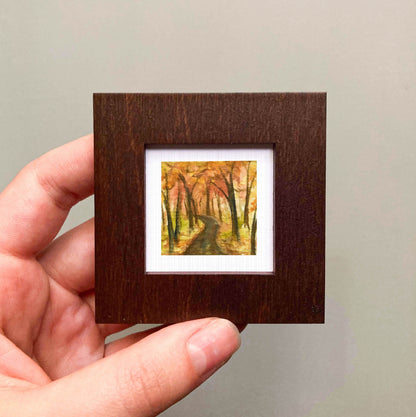 A hand is holding a small wooden frame containing a painting of a forest path with autumn foliage.