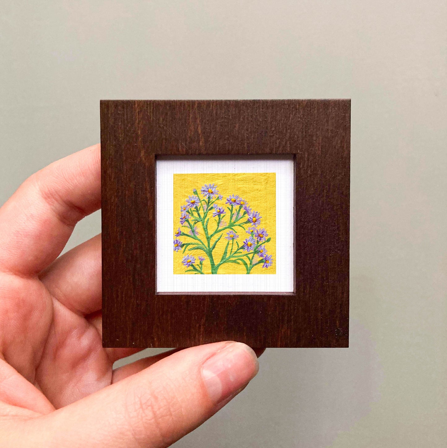A hand is holding a small wooden frame containing a framed painting of a yellow flower with purple petals.