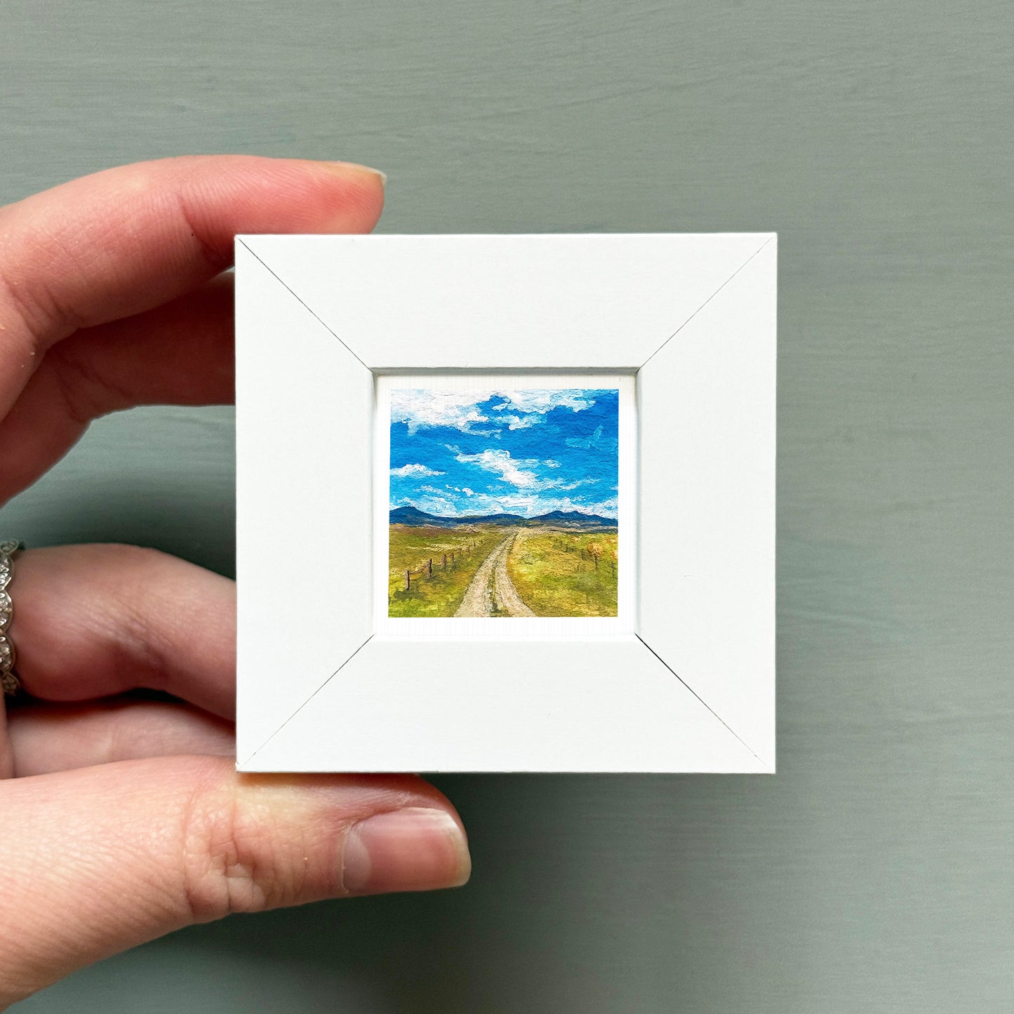 A hand is holding a small white picture frame containing a landscape scene with a dirt road leading to a blue sky with clouds.