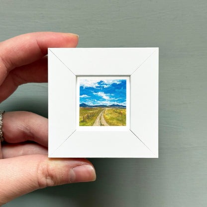 A hand is holding a small white picture frame containing a landscape scene with a dirt road leading to a blue sky with clouds.
