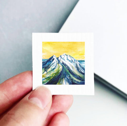 A hand is holding a small square artwork depicting a mountain landscape with a yellow sky.