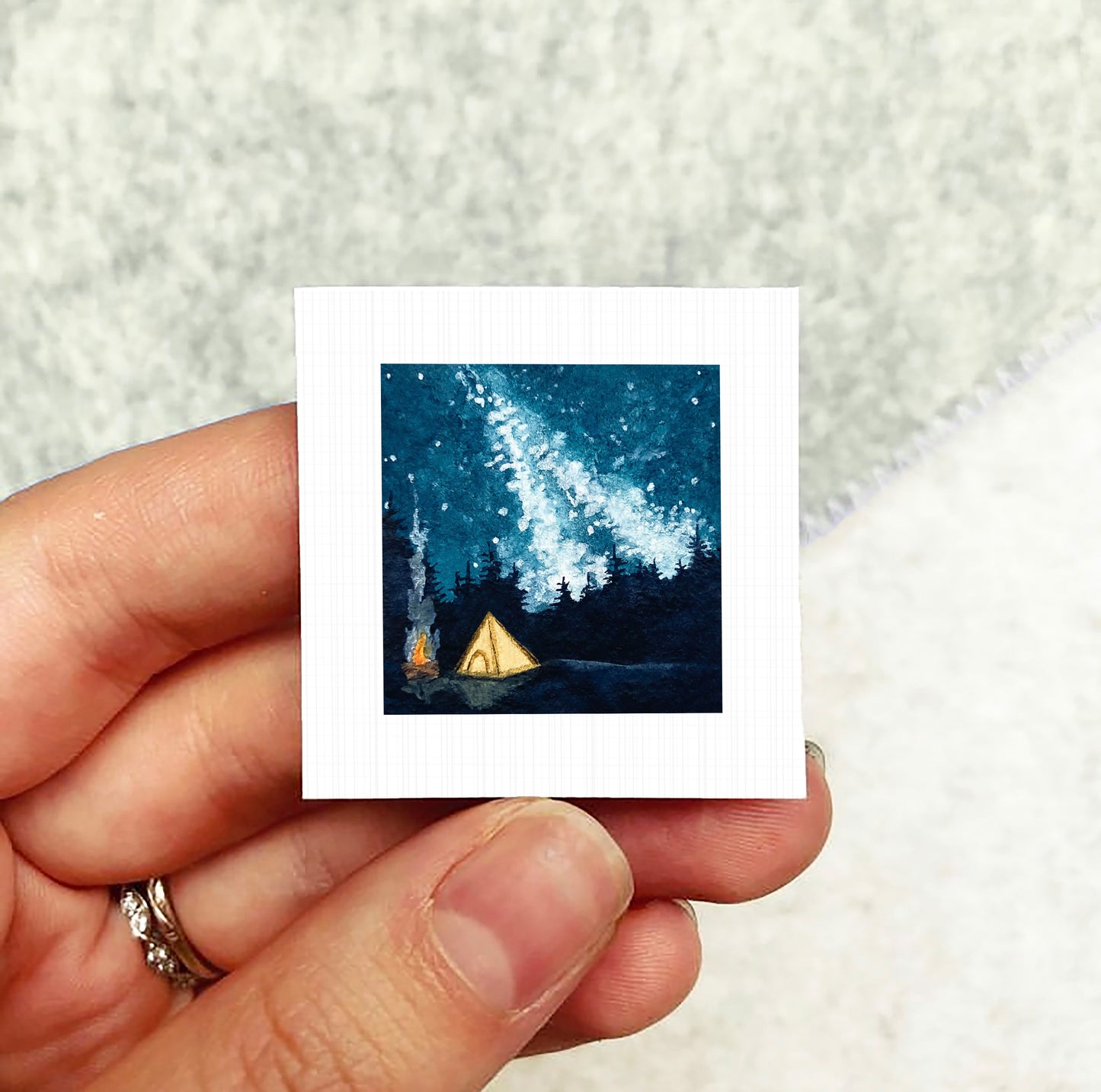 A hand holding a small square artwork depicting a nighttime scene with a tent and a starry sky.