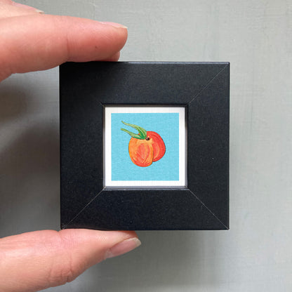 A person's hand holding a small black frame containing a framed illustration of two peaches.