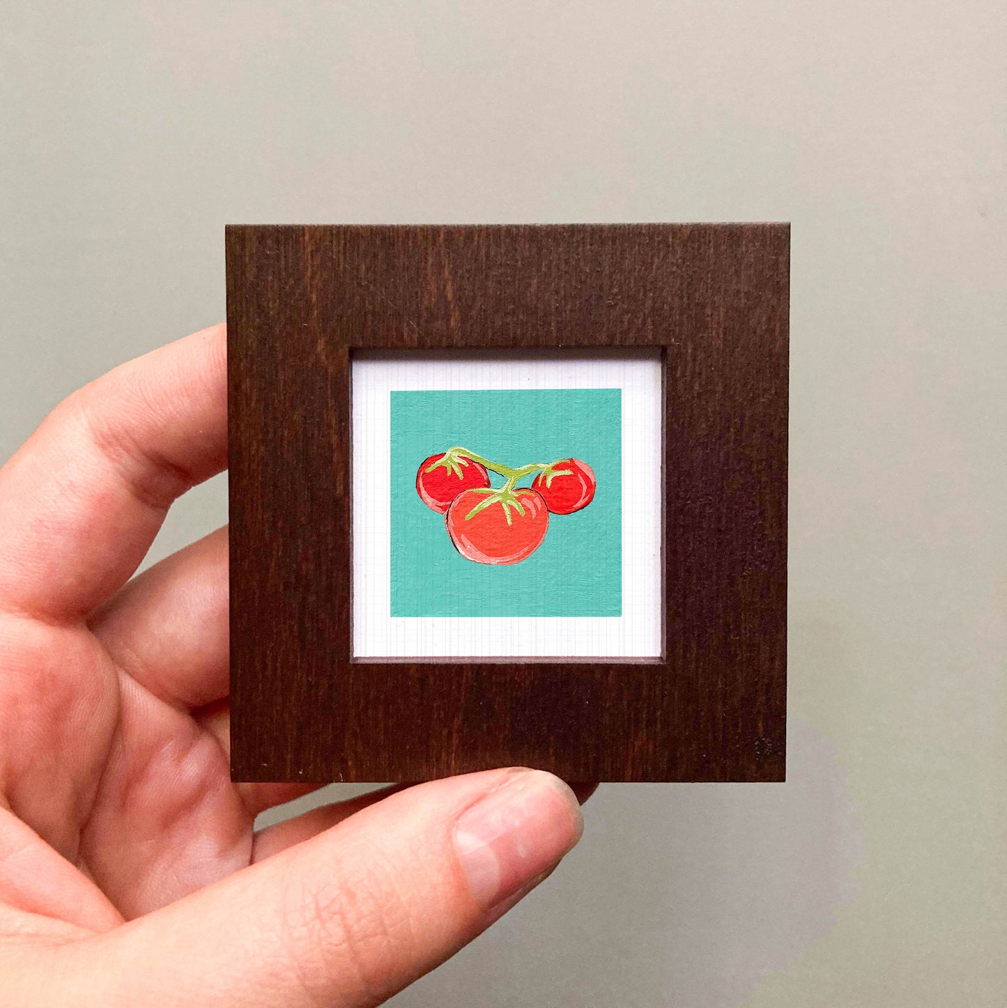 A hand is holding a small wooden frame containing a framed illustration of three tomatoes.