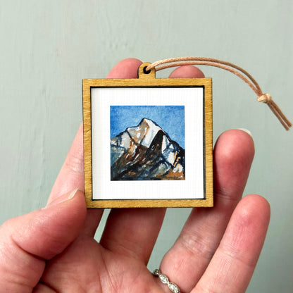 A hand holding a small wooden frame containing a framed painting of a mountain landscape.