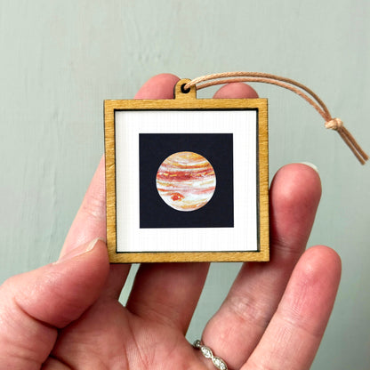 A person's hand holding a small wooden frame containing a framed image of a planet.
