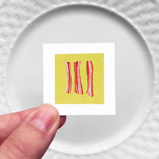 A hand is holding a small square with a picture of three red strips of bacon against a yellow background.
