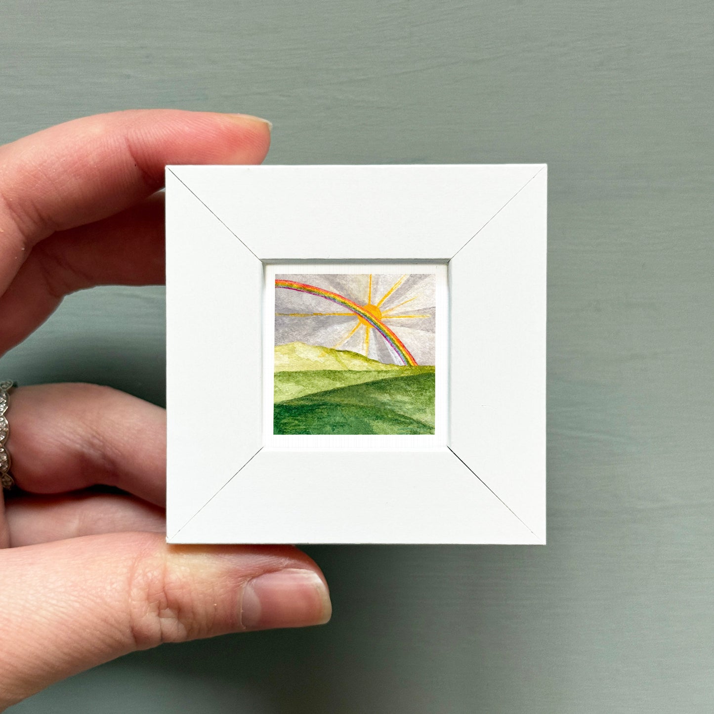 A hand is holding a small white square frame containing a watercolor painting of a landscape with a rainbow in the sky.