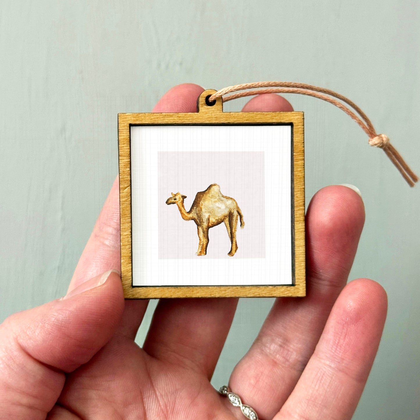 A person's hand holding a small wooden frame containing a picture of a camel.