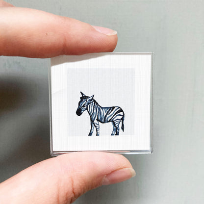 A person's hand is holding a small square with a black and white zebra illustration on it.