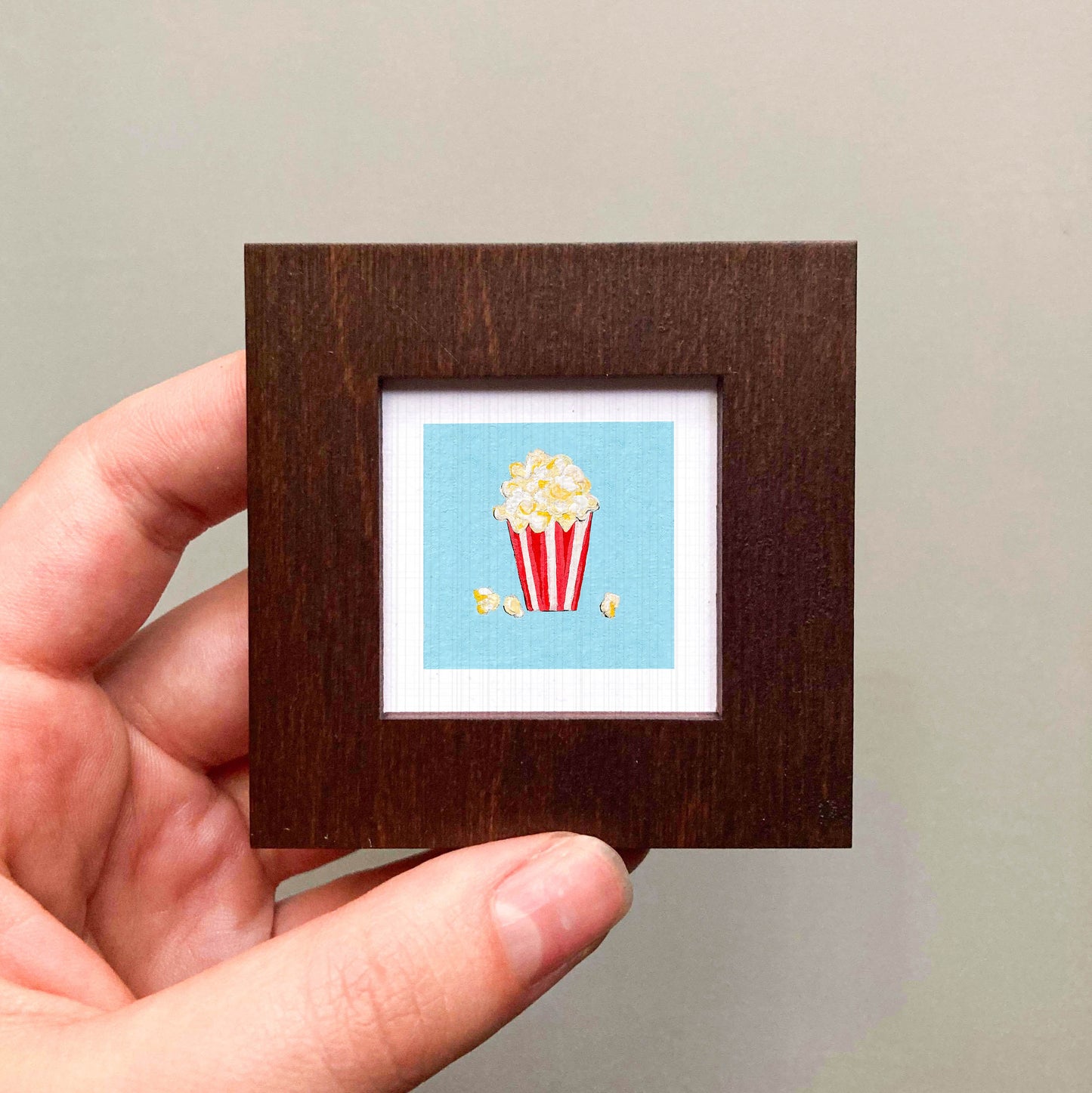 A hand is holding a small square picture frame containing a vibrant illustration of a red and white striped popcorn container.