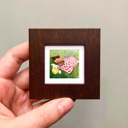 A hand is holding a small wooden frame containing a painting of a picnic scene with a basket, a pitcher, and a red and white checkered blanket on the grass.