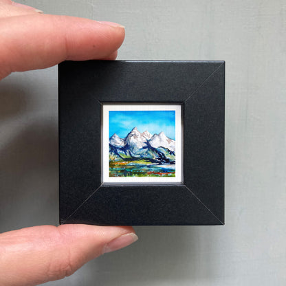 A person's hand holding a small black frame containing a framed landscape painting depicting a mountain scene.