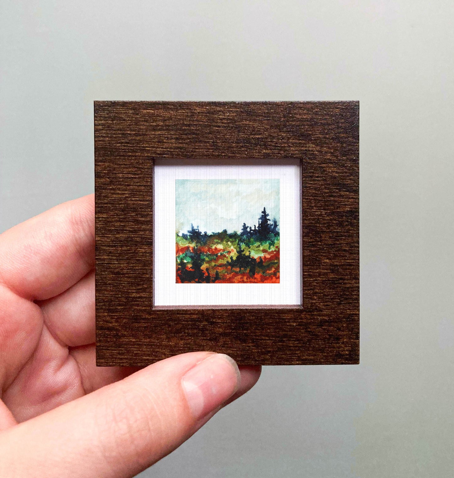 A hand is holding a small, square-shaped wooden frame containing a painting of a landscape with trees and a field of flowers.