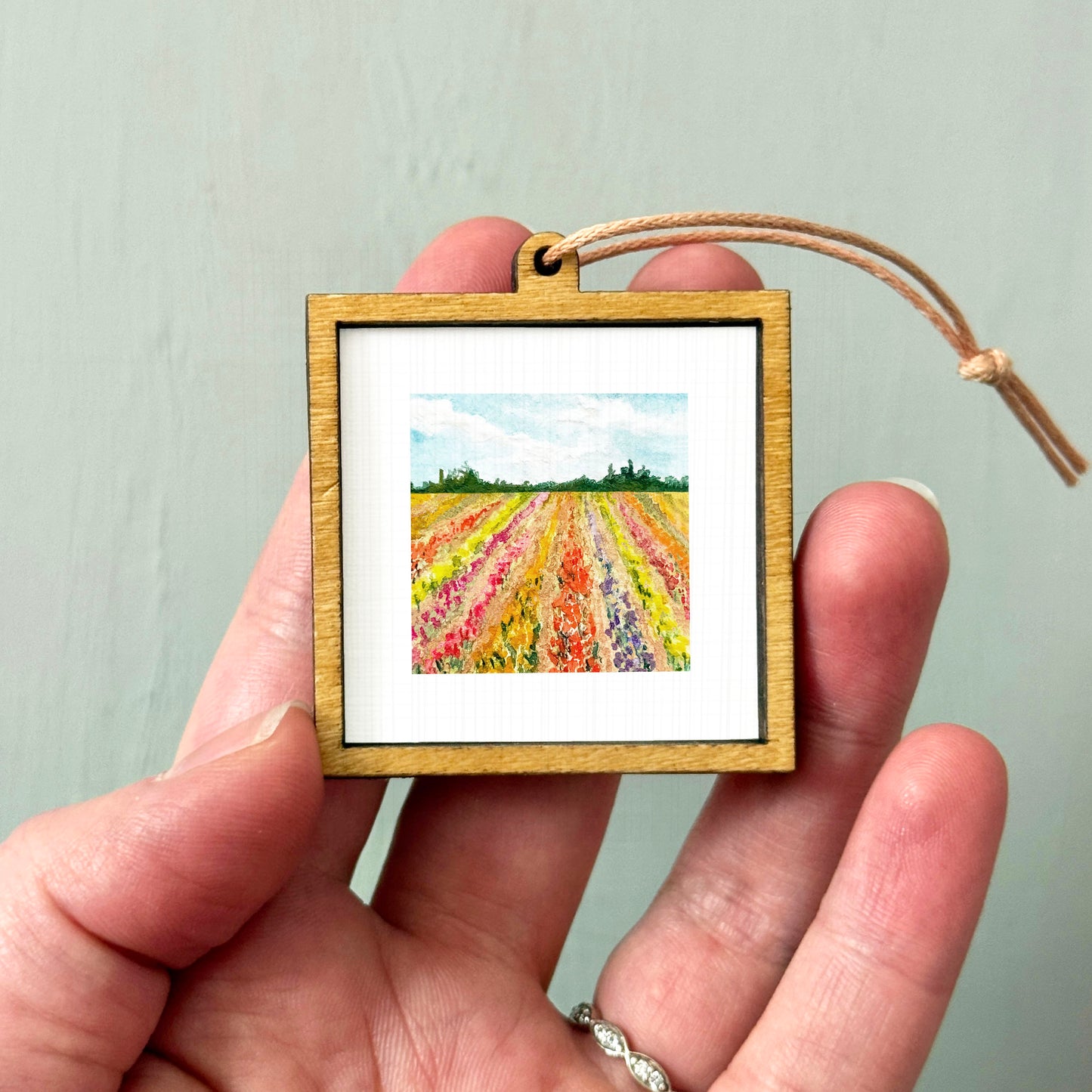 A hand holding a small wooden frame containing a painting of a field with colorful flowers.