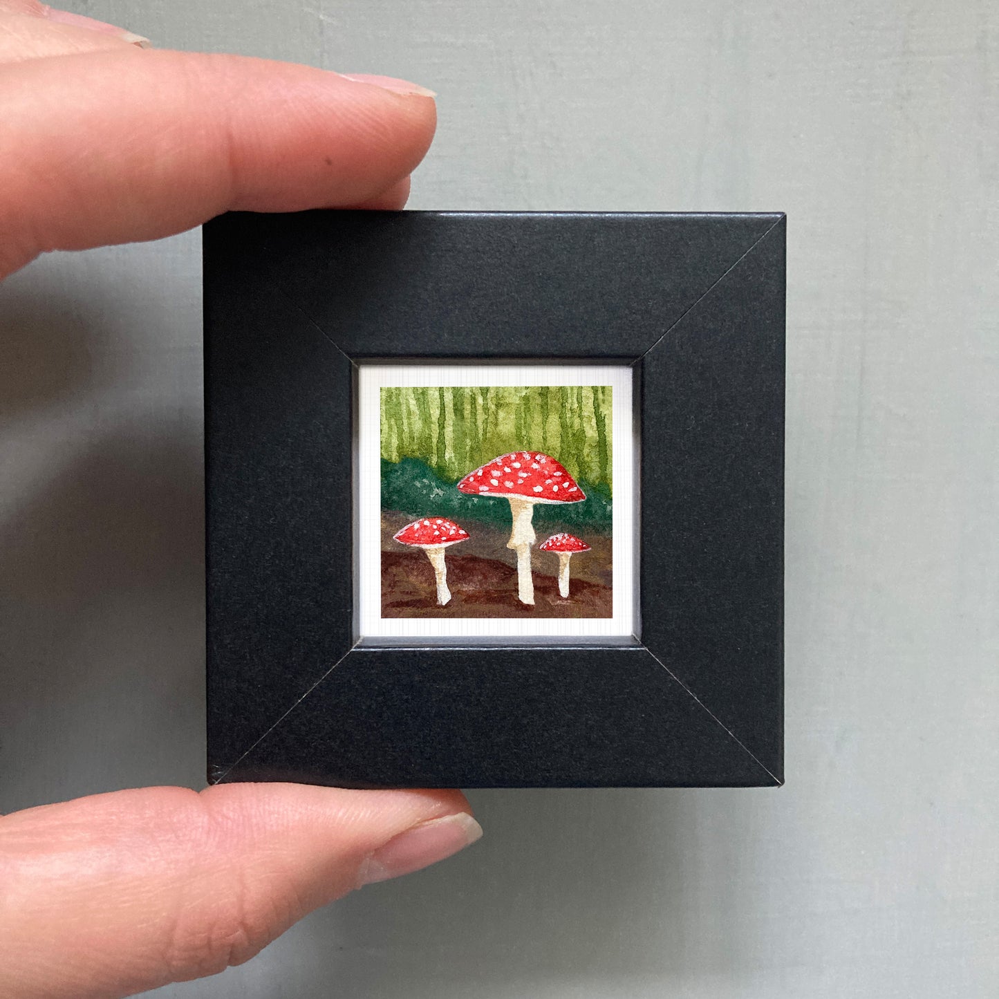 A hand is holding a small black frame containing a watercolor painting of three red mushrooms against a forest background.