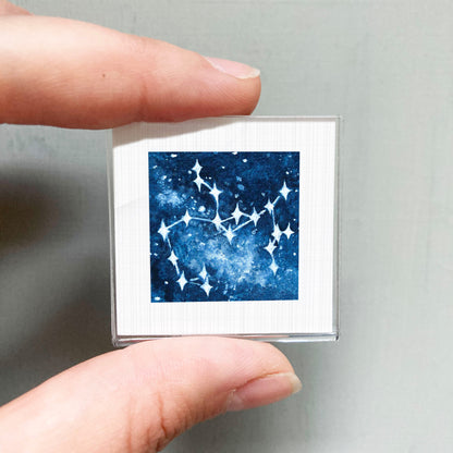 A hand is holding a small square-shaped object with a blue and white starry night sky design on it.
