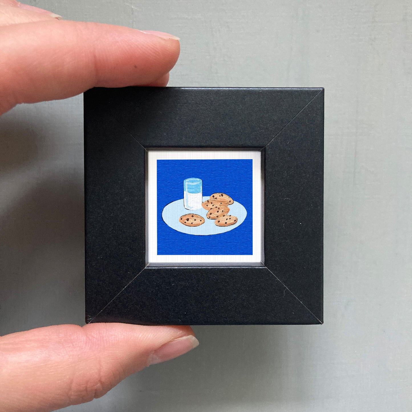 A hand is holding a small black frame containing a framed illustration of cookies and a glass of milk.