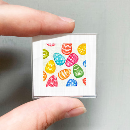 A hand is holding a small square-shaped object with a vibrant pattern of colorful Easter eggs on it.