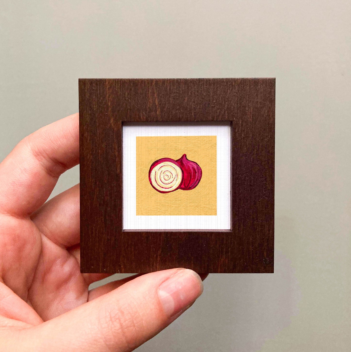 A hand is holding a small wooden frame containing a framed illustration of a red onion.