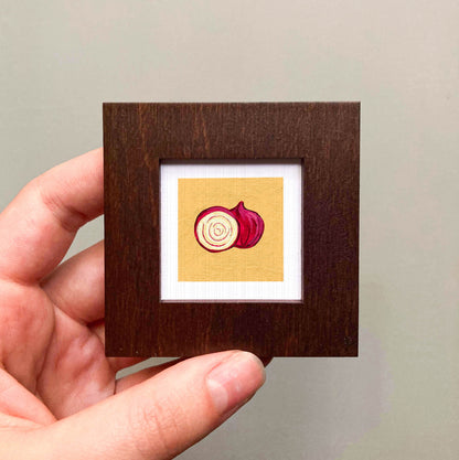 A hand is holding a small wooden frame containing a framed illustration of a red onion.