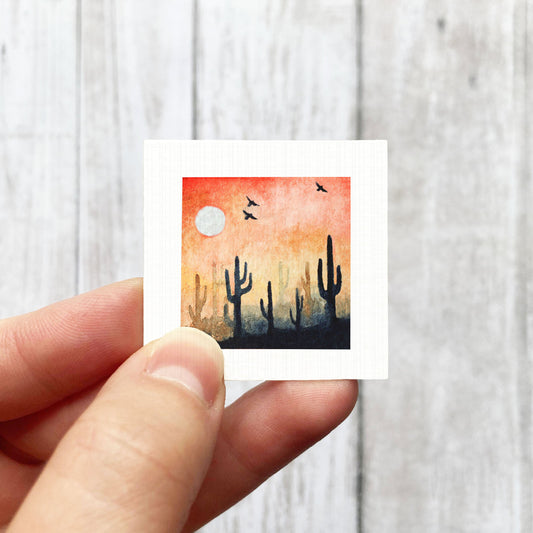 A hand is holding a small square-shaped painting depicting a desert landscape with cacti silhouetted against a colorful sunset sky.