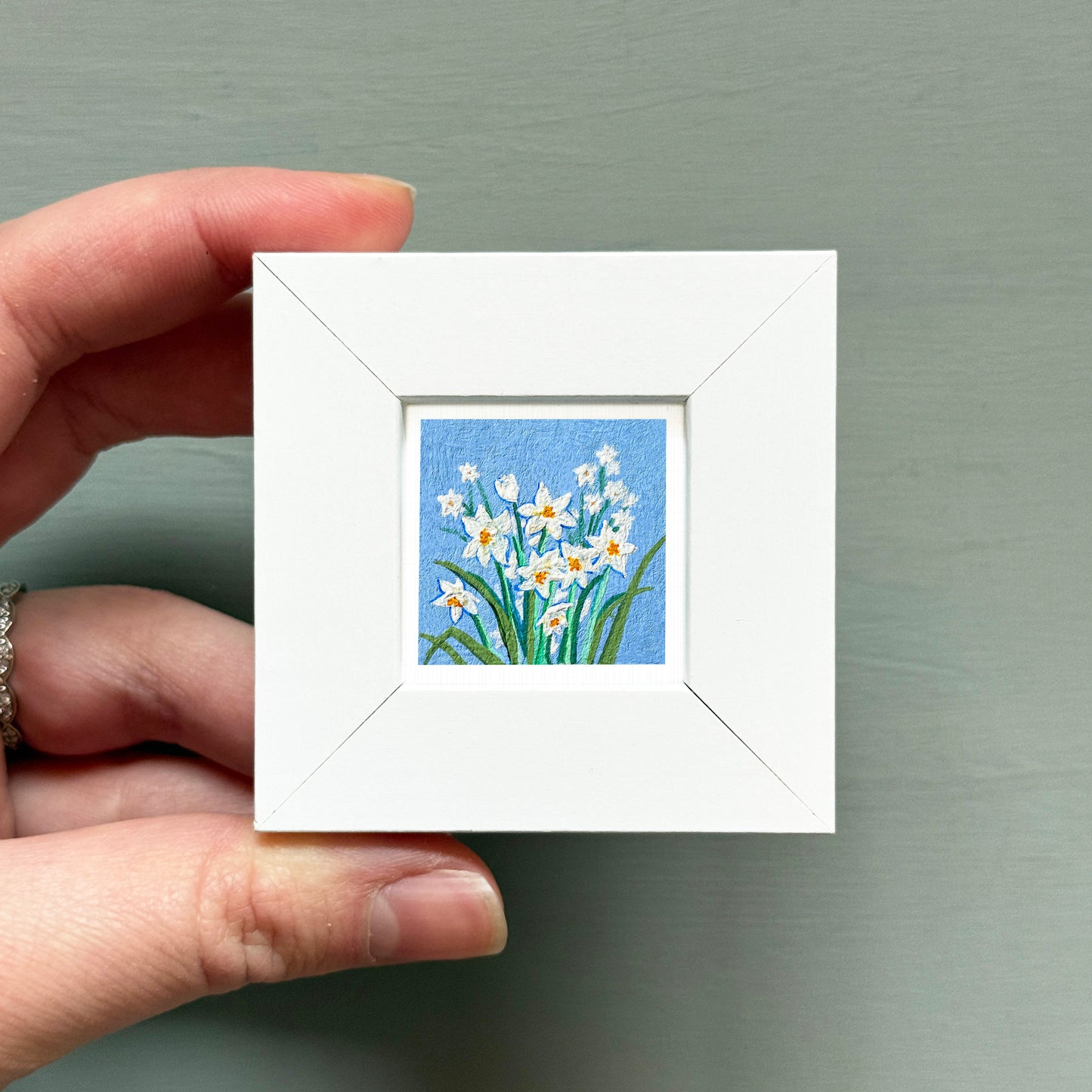 A hand is holding a small white frame containing a painting of white flowers against a blue background.