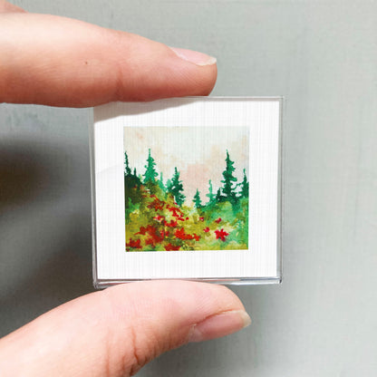 A person's hand holding a small square picture of a forest scene with green trees and red flowers.
