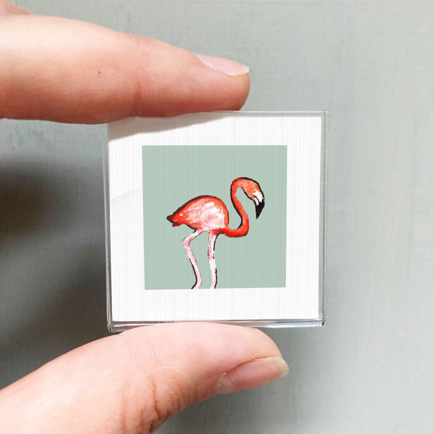 A person's hand is holding a small square picture of a flamingo.