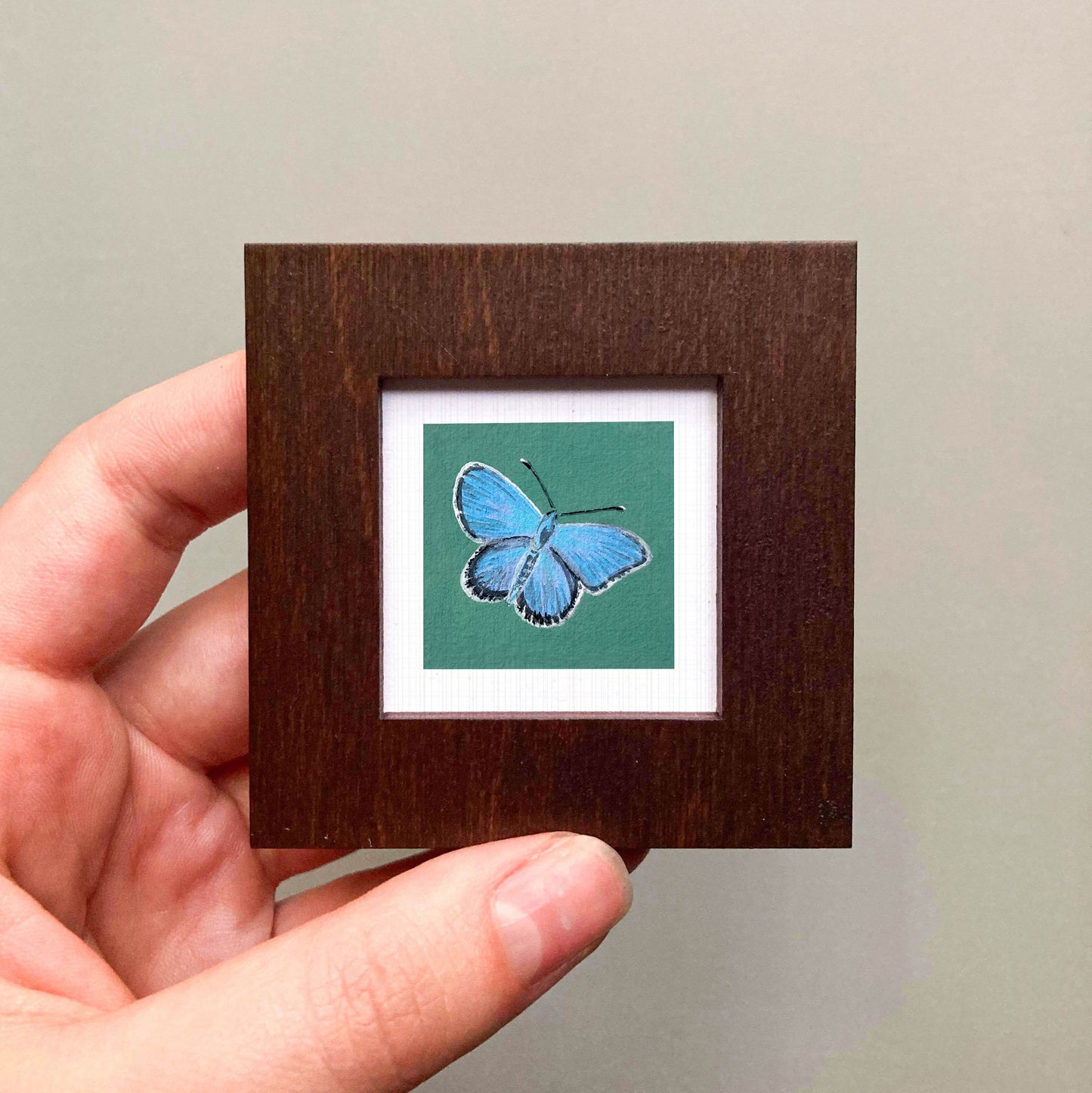 A hand is holding a small wooden frame containing a framed image of a blue butterfly.